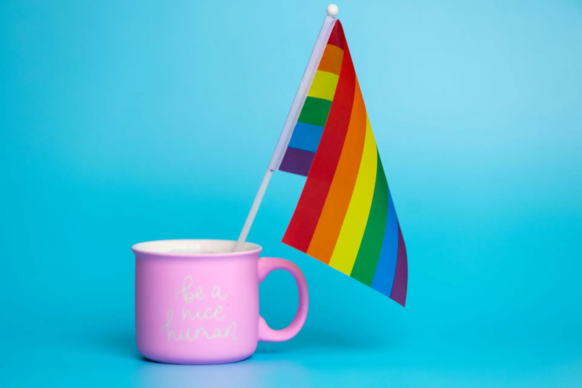 Pink mug with 'be a nice human' text and rainbow pride flag on a blue background.