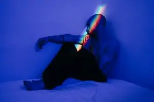 A contemplative shirtless man sits in a blue-lit room, adorned with rainbow light reflections.