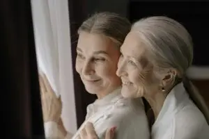 Senior women couple smiling and embracing while looking out a window, showcasing love and happiness.